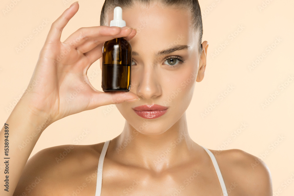 Fototapeta premium Beautiful young woman holding amber glass dropper bottle near face, showcasing skincare product on beige background.