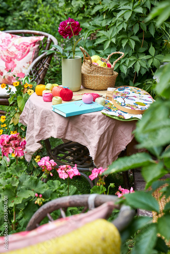 A small table covered with a soft pink cloth holds embroidery threads, a piece of fabric with an embroidery design, a notebook, and a glass vase with a blooming garden peony. 