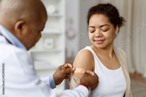 Doctor applying adhesive bandage to patients upper arm after vaccination, smiling african american woman got injection in bright medical room, had immunization treatment in modern clinic
