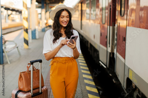 Young attractive lady traveler standing with suitcase at railway station platform with cellphone in hands, checking ticket and waiting for departure