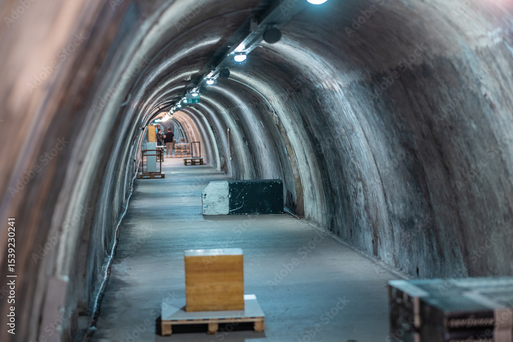 Fototapeta premium tunnels under Zagreb, featuring a curved, concrete passageway with dim lighting from overhead lamps. Scattered crates and pallets suggest storage or construction, while a few people are visible in the