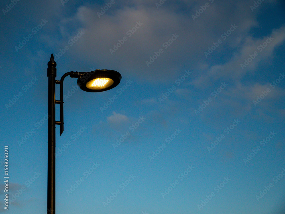 A street LED light is lit up in the evening sky. The sky is mostly clear with a few clouds scattered around. The light is shining brightly, illuminating the area below. The scene has a peaceful mood A street LED light is lit up in the evening sky. The sky is mostly clear with a few clouds scattered around. The light is shining brightly, illuminating the area below. The scene has a peaceful mood