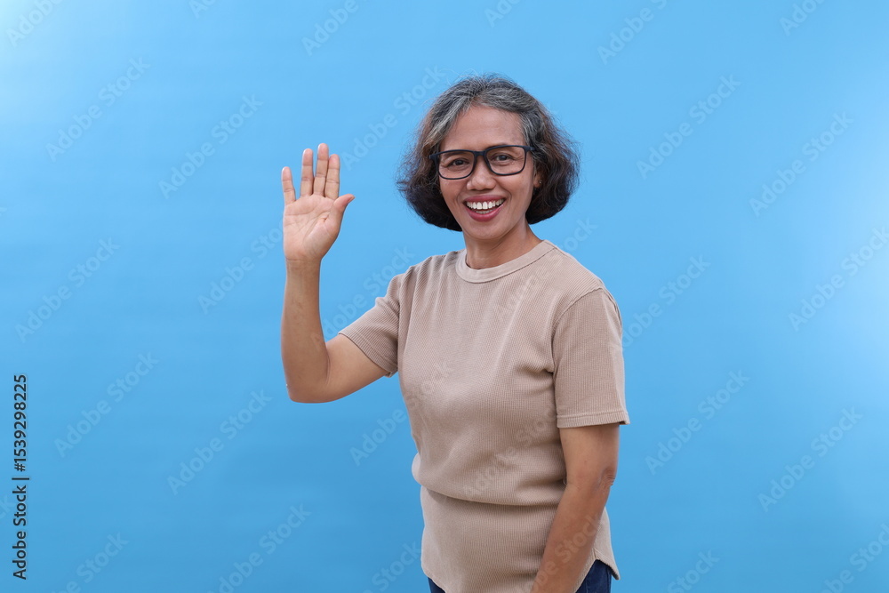 Fototapeta premium Gray-haired Asian old woman, wearing glasses, waving hand, greeting someone in a friendly manner, looking at camera with smile, standing isolated on blue background.