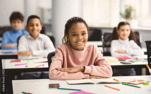 Elementary Education. Portrait of happy small african american girl sitting at table in classroom at primary school with group of classmates and posing, looking at camera and smiling