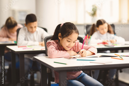 Education And Studying Concept. Portrait of smiling asian small student sitting at desk in classroom, writing in notebook or drawing picture with colorful pencils. Back To School