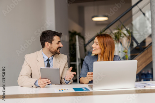 Two executives smiling at each other at corporate office and using laptop and tablet for case study.