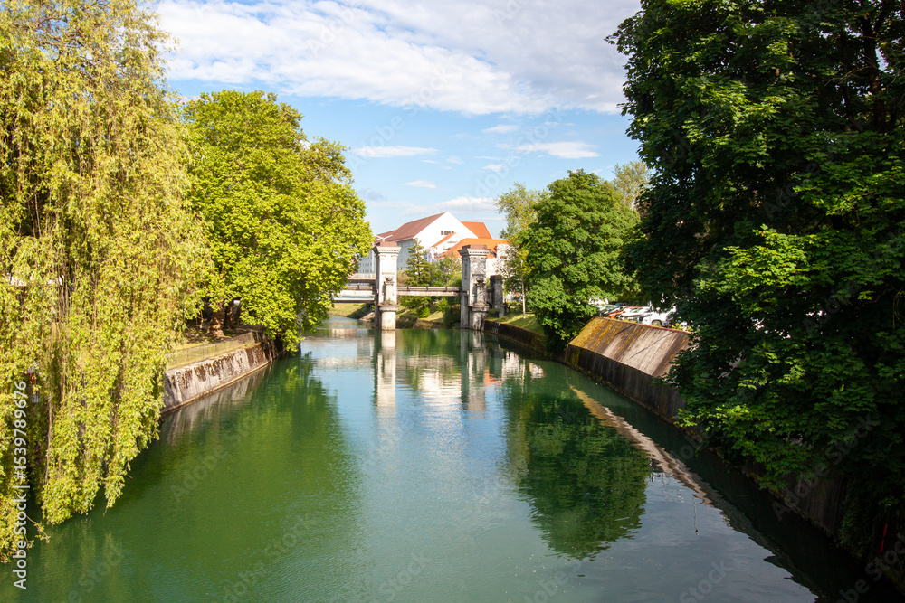 Obraz premium A serene and picturesque view of a tranquil canal or river in Ljubljana, on a bright, sunny day with a clear blue sky dotted with fluffy white clouds