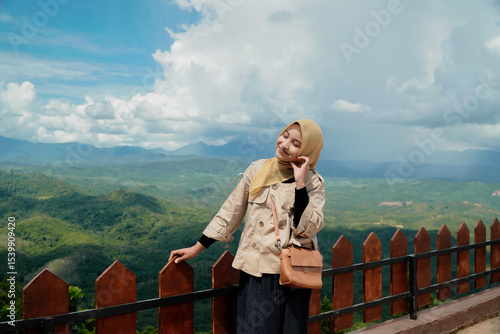 Woman in hijab enjoys scenic mountain vista, leaning on wooden fence.
