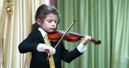 A little boy in a suit and festive tie plays the violin at a music school concert, trying hard and expressing his emotions.