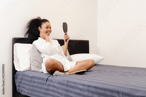 Foto Beautiful curly hair woman in white bathrobe hold mirror, trying to improve her appearance while sit on bed in bedroom