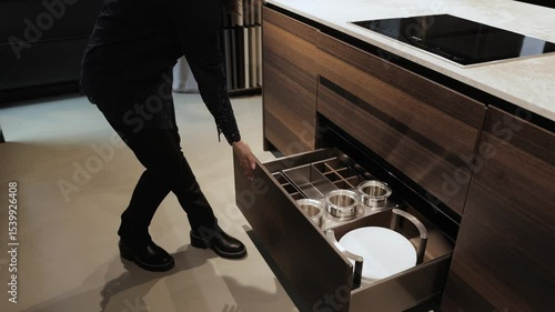 Woman opens drawers of dark modern kitchen cabinets and shows dishes inside. Kitchen island, modern kitchen interior design.