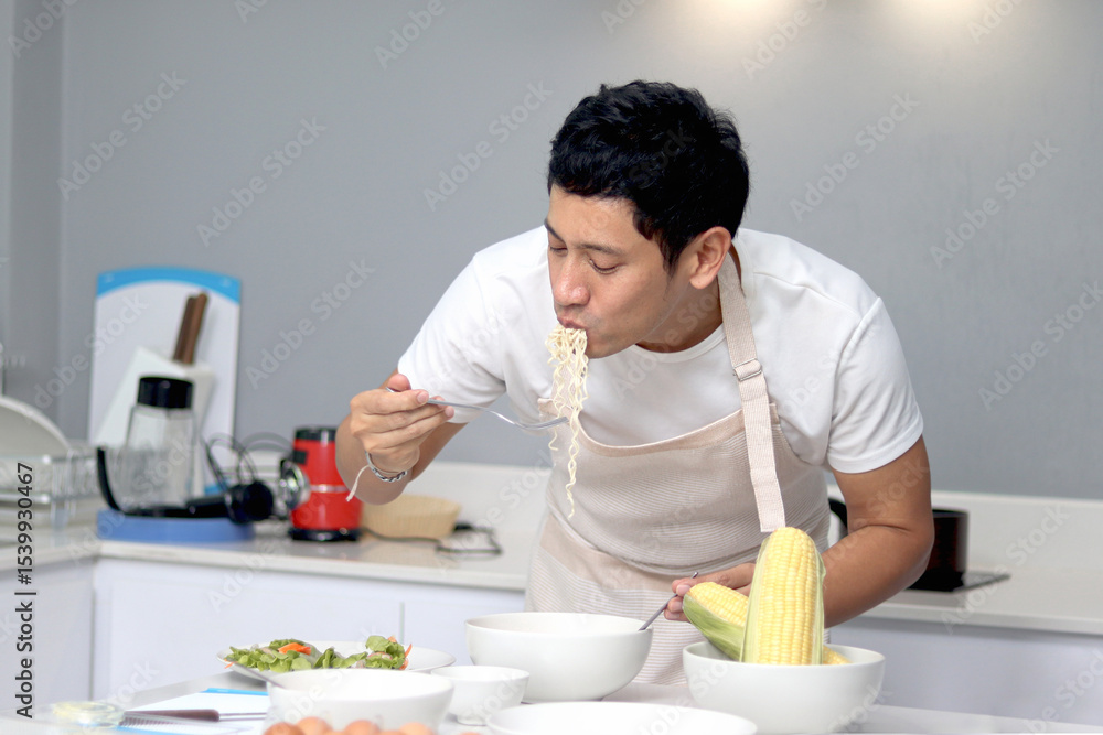 Asian man with apron eating noodle after preparing breakfast meal in the kitchen. Joyful man cooking food and eating meal in kitchen at home in the morning. A man is eating delicious noodle. Asian man with apron eating noodle after preparing breakfast meal in the kitchen. Joyful man cooking food and eating meal in kitchen at home in the morning. A man is eating delicious noodle.