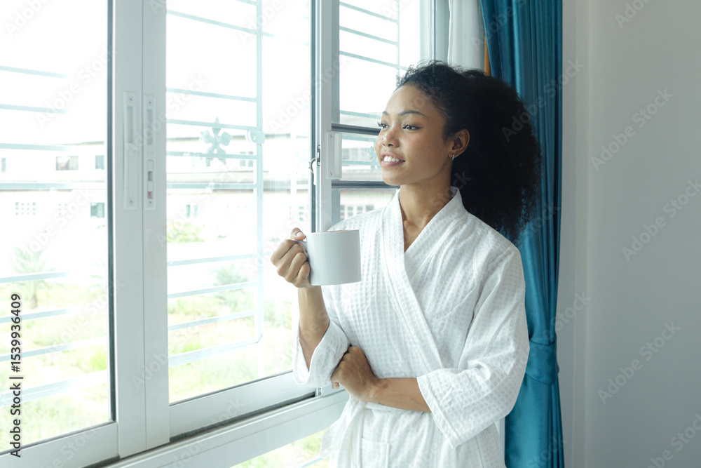 Beautiful curly hair woman in white bathrobe holding coffee mug while standing in the bedroom after waking up in morning. Happy African female drinking coffee, feeling refreshed after waking up. Beautiful curly hair woman in white bathrobe holding coffee mug while standing in the bedroom after waking up in morning. Happy African female drinking coffee, feeling refreshed after waking up.