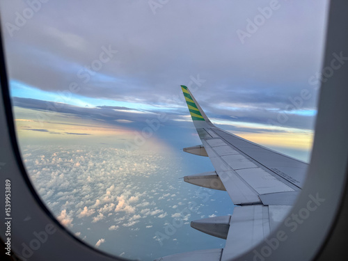 Wing view from an airplane window during flight, showcasing a serene sky with scattered clouds below and a soft rainbow glow on the horizon at golden hour.