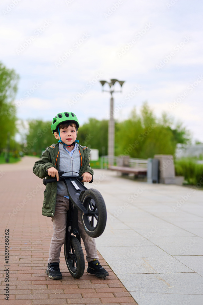 Obraz premium boy with green helmet holding balance bike in park. child learning cycling skills, outdoor activity, healthy lifestyle concept. bright day, trees and benches in background