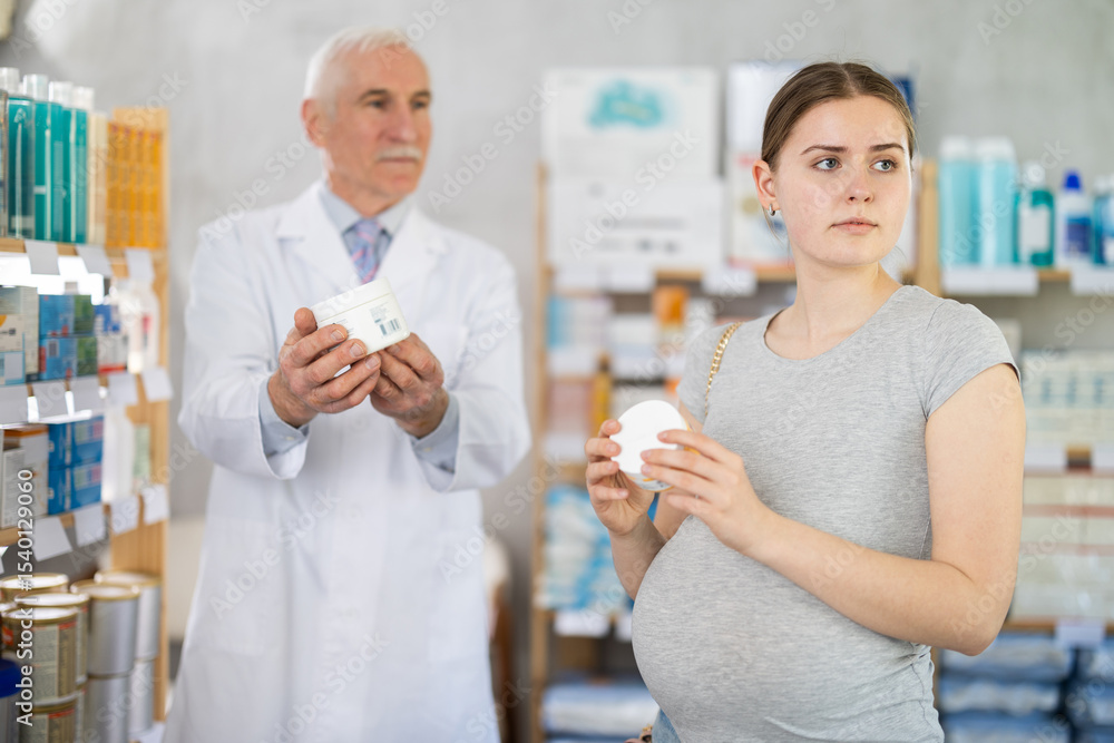 Obraz premium Pregnant girl looking for skin cream in a pharmacy. Male pharmacist helps to choose right skin cream
