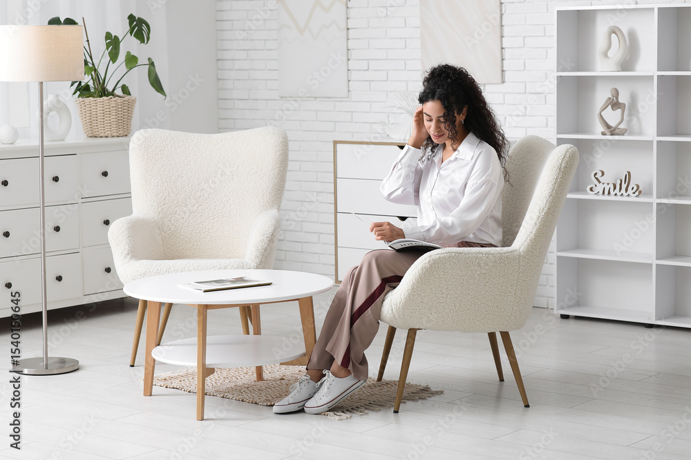 Pretty young African-American woman in stylish white shirt with magazine sitting on armchair at home Pretty young African-American woman in stylish white shirt with magazine sitting on armchair at home