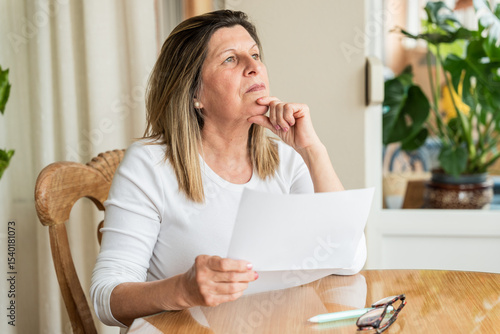Fotografie Senior woman sitting at dining table, holding documents and looking pensive, con