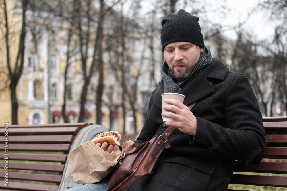 Pensive hipster drinking coffee sitting on a bench in the urban park. Handsome man wearing black coat eating street food, hot dog with paper cup in hand in the city. Stylish bearded middle-aged male. Pensive hipster drinking coffee sitting on a bench in the urban park. Handsome man wearing black coat eating street food, hot dog with paper cup in hand in the city. Stylish bearded middle-aged male.