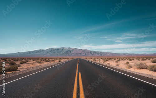 A straight asphalt road in the desert leads toward a distant mountain range under a bright blue sky with soft clouds.