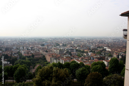 Hazy, elevated view of Bergamo's cityscape with red roofs and numerous trees. Muted atmosphere