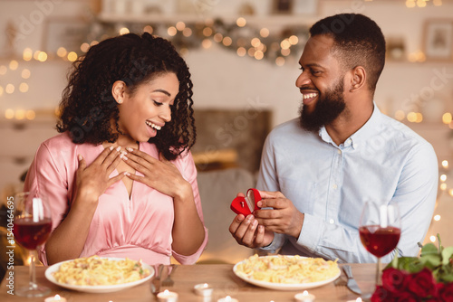 Will You Marry Me. Handsome bearded african american man holding and showing open box, offering marriage ring to his happy fiance, getting engaged in restaurant or at home during romantic date