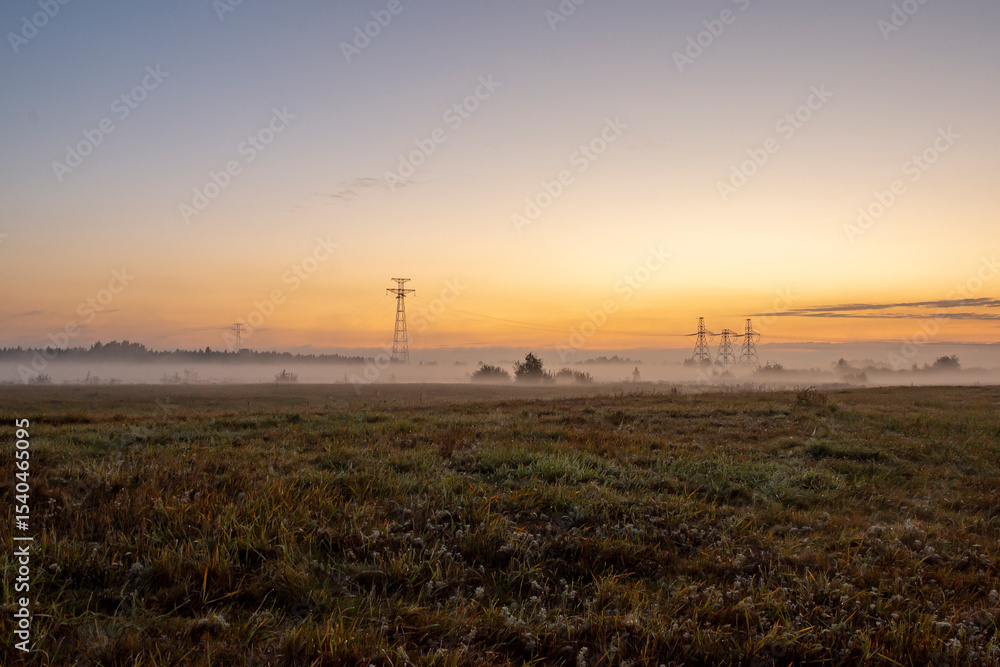 Obraz premium The morning light illuminates a pastoral view where a power transmission tower rises among autumn meadows and fields.