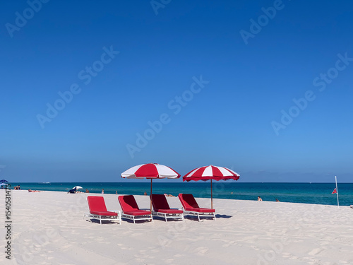 Bright red beach umbrellas and lounge chairs sit on a pristine white sand beach with a vivid blue sky and turquoise ocean in the background, creating a vibrant tropical vacation scene.