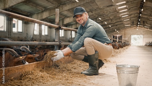 Good-looking hard working man with gloves holding hay and feeding goats and lambs in shed. Attractive worker smiling, happily looking at camera. Concept of farming, village lifestyle.