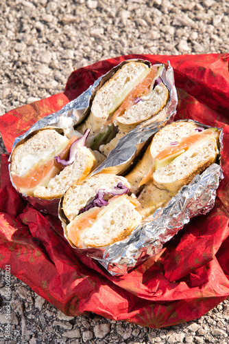 Two halves of freshly made bagels filled with smoked salmon, cream cheese, cucumbers, and red onions, wrapped in aluminum foil and placed on decorative red paper, creating a tempting takeaway meal