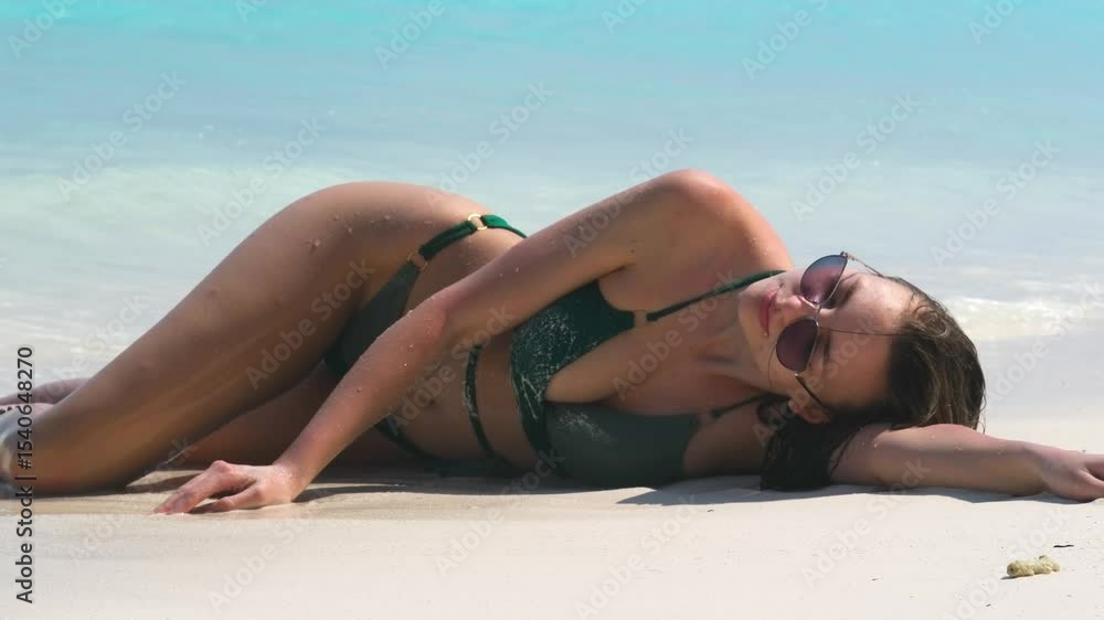 Young woman in green bikini posing sensually on sandy beach, laying in water with relaxed expression, turquoise ocean in background, summer vacation, travel, tourism, summer tourist relaxing Young woman in green bikini posing sensually on sandy beach, laying in water with relaxed expression, turquoise ocean in background, summer vacation, travel, tourism, summer tourist relaxing