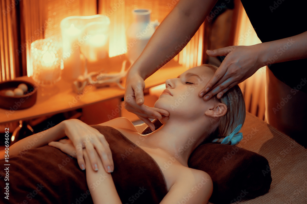 Fototapeta premium Caucasian woman enjoying relaxing anti-stress head massage and pampering facial beauty skin recreation leisure in warm candle lighting ambient salon spa in luxury resort or hotel. Quiescent