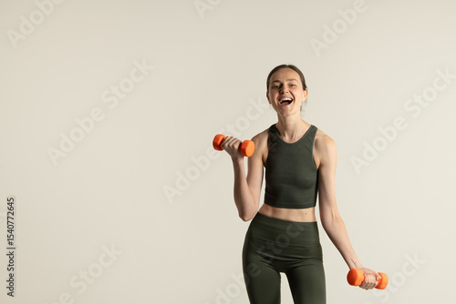 A girl wearing a green form-fitting outfit joyfully lifts orange dumbbells, showcasing her commitment to fitness in a simple yet stylish indoor setting during daytime