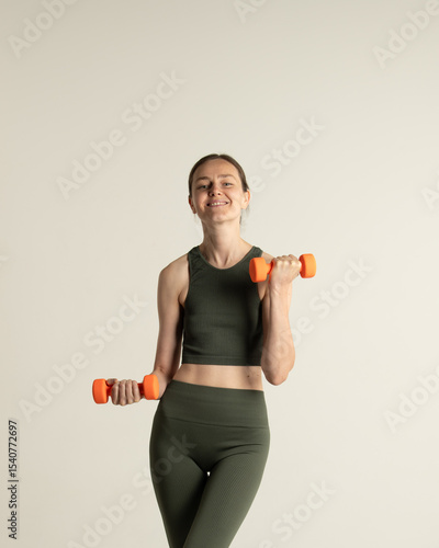 A girl dressed in a green crop top and leggings is exercising indoors, holding orange dumbbells. She appears happy and engaged in her fitness routine, showcasing her workout enthusiasm
