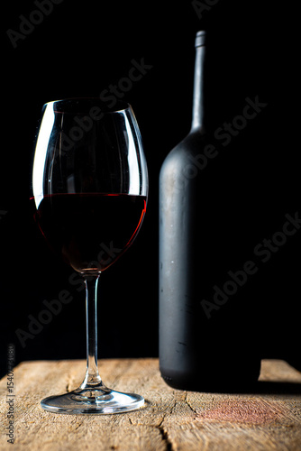 Obraz na plátně Taça de cristal com vinho tinto e garrafa sobre a mesa de madeira rústica, com fundo preto