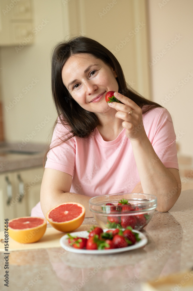 Smiling woman in a kitchen with fresh fruit.. Embracing playful skincare with fresh strawberry slices and a big smile Smiling woman in a kitchen with fresh fruit.. Embracing playful skincare with fresh strawberry slices and a big smile