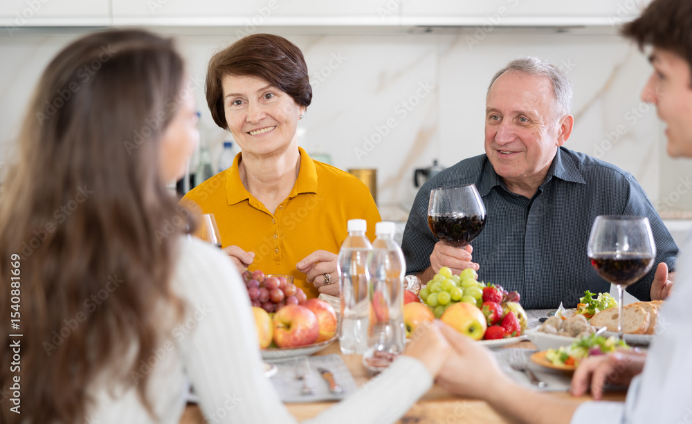 Happy carefree senior parents sharing memories with grown daughter and son-in-law during conversation over family celebration with wine at cozy home environment Happy carefree senior parents sharing memories with grown daughter and son-in-law during conversation over family celebration with wine at cozy home environment