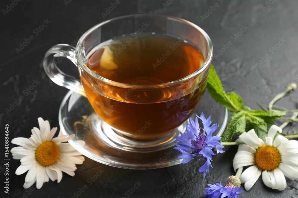 Aromatic herbal tea in glass cup, different flowers and mint on black table, closeup Aromatic herbal tea in glass cup, different flowers and mint on black table, closeup