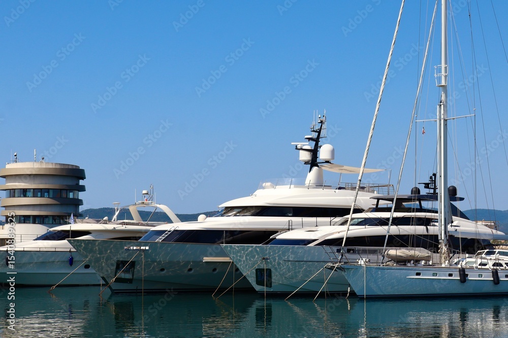 Fototapeta premium Luxury yachts are moored in a calm marina under a clear blue sky. The reflections of the boats shimmer on the water. A modern round harbor building is visible in the background.