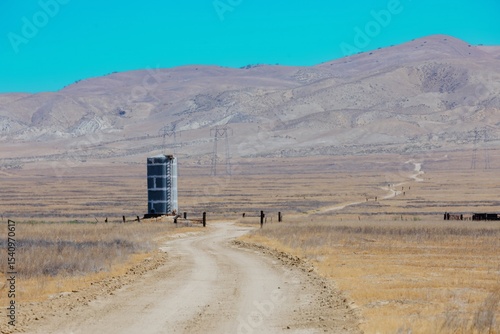 A dirt road leads to a water tank in a dry, grassy field in Carrizo Plain National Monument, California, USA . The tank provides water for cattle grazing in the area. Mountains are in the background.