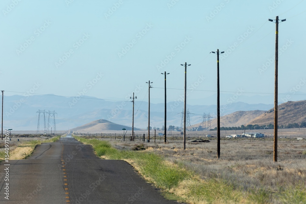 Fototapeta premium A desolate road stretches through arid landscape in Carrizo Plain National Monument, California, USA. Power lines run alongside, connecting distant towers, symbolizing energy transport.