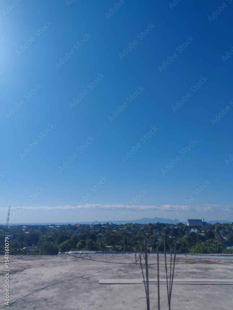 Fototapeta premium Open construction site with exposed rebars and panoramic view of a tropical town under a clear blue sky and distant mountain range