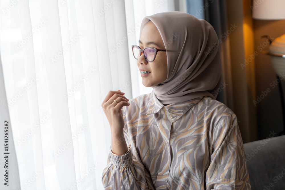 Obraz premium Asian Indonesian Muslim Woman with Hijab Looking Out Window Indoors