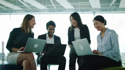 A diverse group of young professionals laugh and collaborate with laptops in a bright, modern office, showcasing teamwork and positive workplace culture.