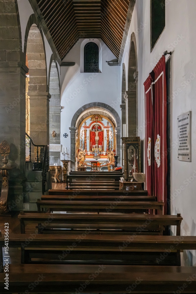 Fototapeta premium Inside a historic Portuguese church, featuring chandeliers hanging from the high ceiling, leading towards the illuminated altar. The nave is lined with traditional wooden pews and stone columns