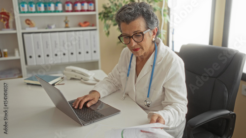 Wallpaper Mural Senior woman doctor with grey hair working in office clinic on laptop wearing stethoscope surrounded by medical files and telephone in a professional healthcare environment. Torontodigital.ca