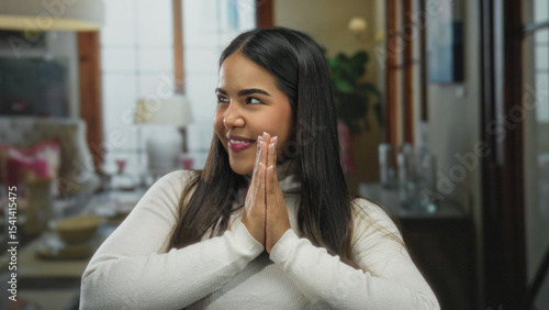 Hispanic woman smiling contently with hands together indoors  surrounded by a cozy room setting  exuding peace and warmth during a serene moment of meditation.