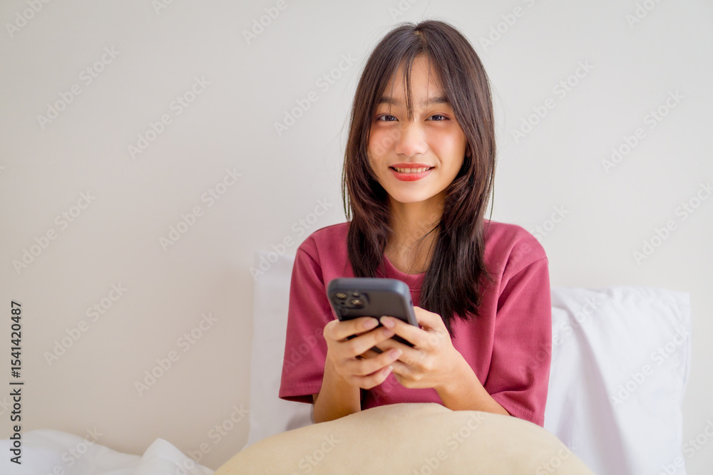 Asian young girl using smartphone texting and chatting with friend in bedroom at home. Relaxing woman wearing pink shirt sit on the bed using mobile phone to communicate in the morning at house. Asian young girl using smartphone texting and chatting with friend in bedroom at home. Relaxing woman wearing pink shirt sit on the bed using mobile phone to communicate in the morning at house.