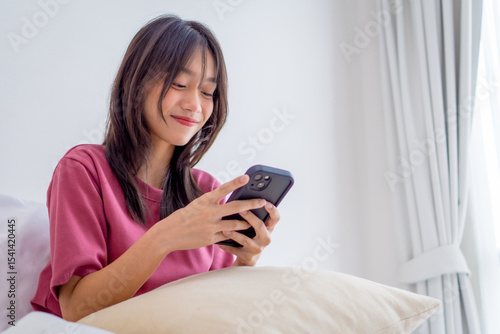 Asian young girl using smartphone texting and chatting with friend in bedroom at home. Relaxing woman wearing pink shirt sit on the bed using mobile phone to communicate in the morning at house.