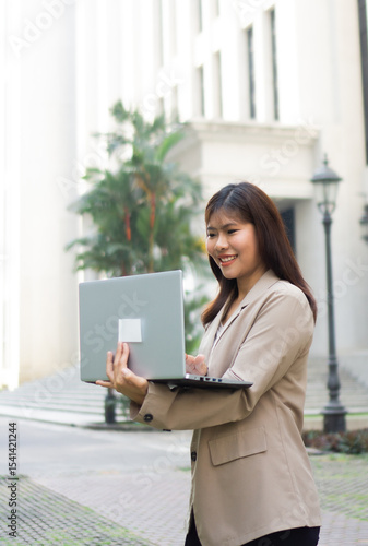 Young happy Asian businesswoman working on laptop in city. Confident female executive using computer while standing outdoors in urban business district, multitasking and managing work on the go.